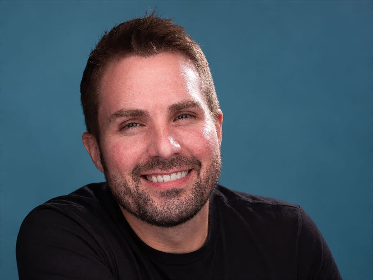 Scott Squire smiling, in a black T-shirt against a blue backdrop